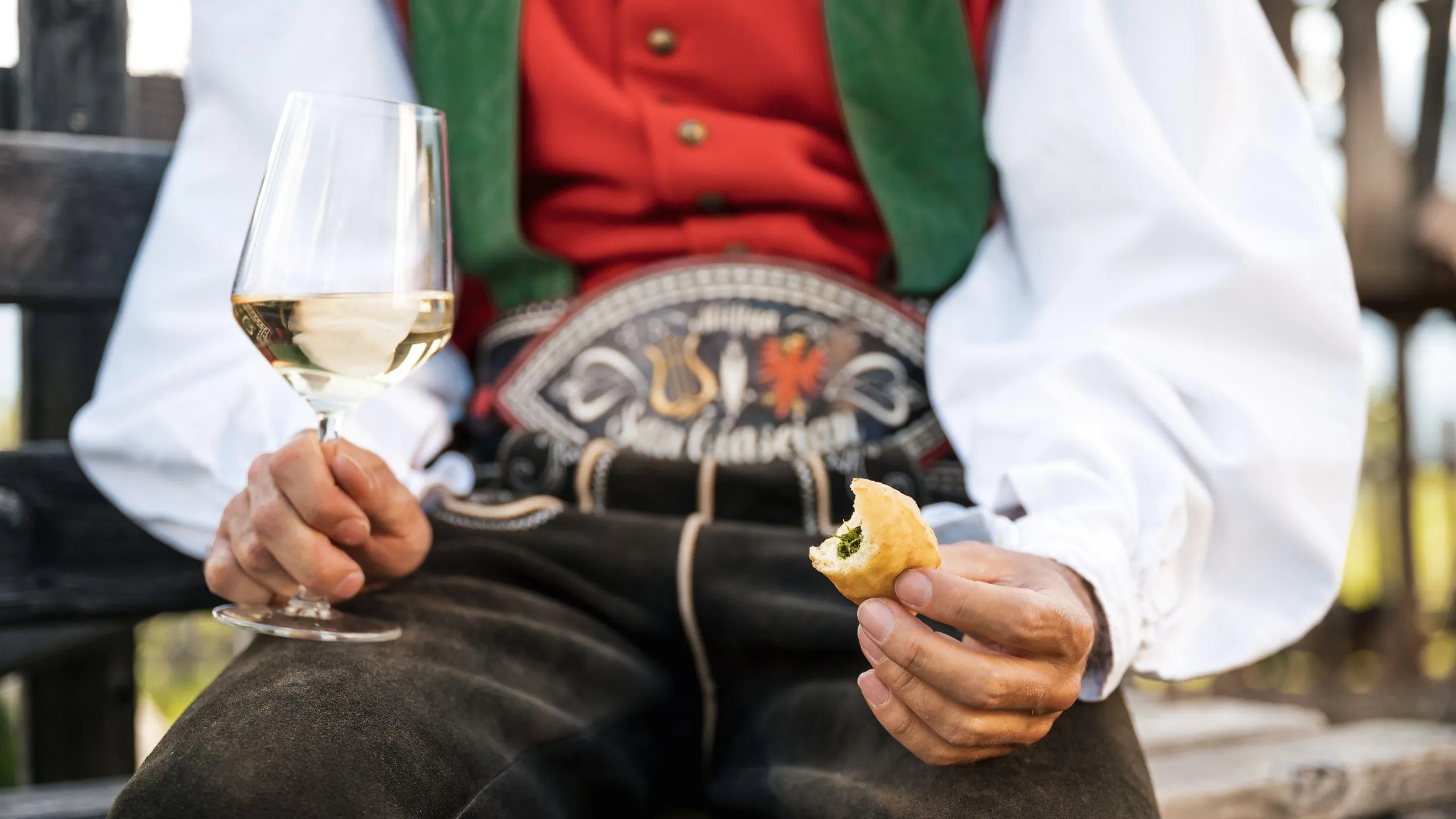 A person in traditional attire holds a glass of white wine in one hand and a Cascencel ArstĂ in the other while seated on a wooden bench.