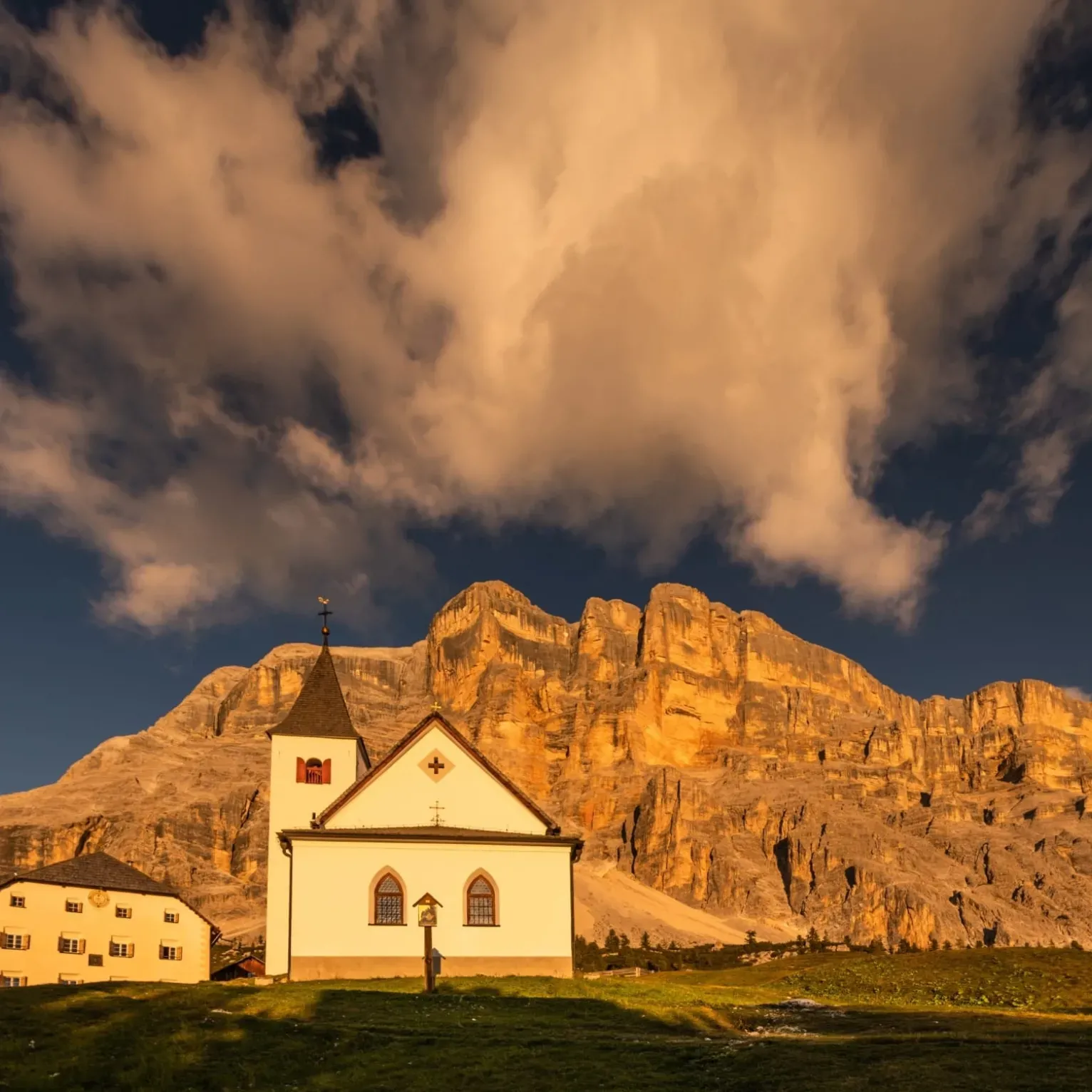 The small white church of La Crusc/Santa Croce in Alta Badia with a steep roof stands in front of the majestic mountain range of Sas dla Crusc/Sasso Croce under a dramatic sky filled with clouds.