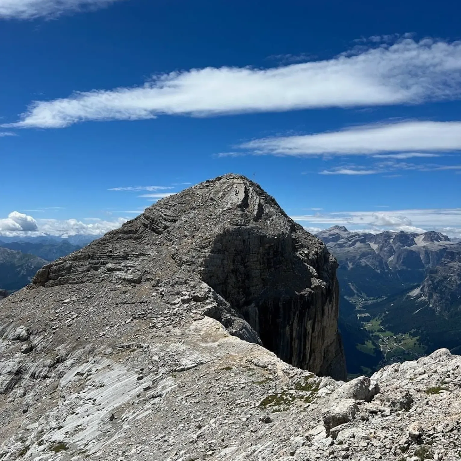 The peak of the Ciaval-Sas dla Crusc rises against a blue sky with scattered clouds, overlooking a valley with green landscapes and distant mountain ranges.
